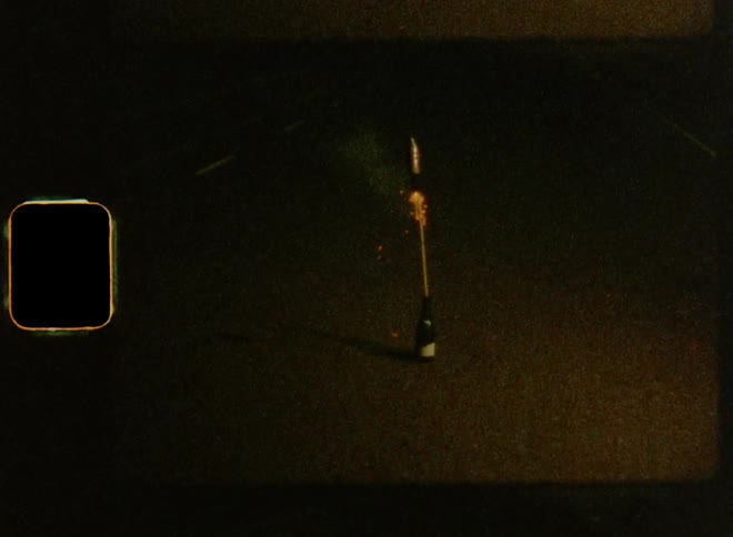 A young Caucasian man kneels in the street as he lights a bottle rocket and it takes off into the dark sky on New Year’s Eve on Super-8 mm film