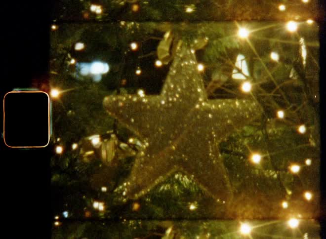 Hand-held retro Super-8 film footage close-up of reflective and glittering sparkling star on decorated Christmas tree with lights of light chain causing lens flares during advent time or nighttime on X-mas evening with festive atmosphere and cosy light