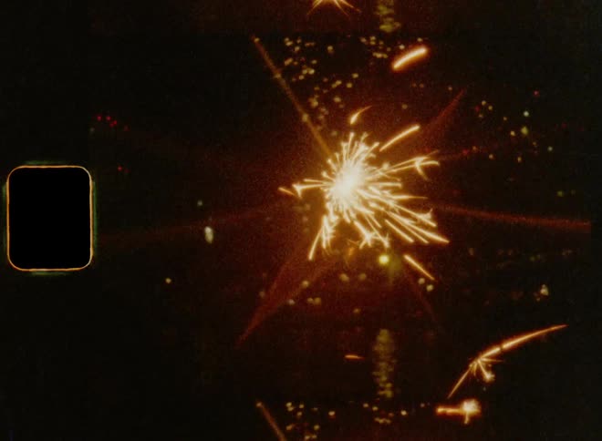 Close up of a sparkler in front of a fireworks show in the night sky caught on vintage film