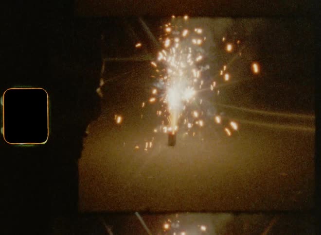 The silhouette of two friends standing outdoors facing a fireworks sparkler going off on the ground on Super-8 mm film