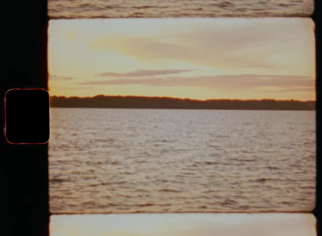 Ripples cover the surface of a large body of water that is washing upon a coastal shoreline as the sun makes the red clouds lighten up the sky during the last daylight on a warm summer evening by the sea on retro 8mm film