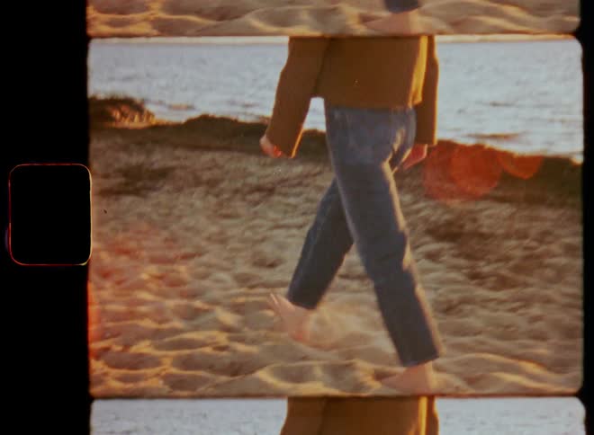 Young woman wearing retro jeans walking barefoot over the warm sand on a beach by the sea at sunset on vintage 8mm film with lens flare and stylish fashion vibes