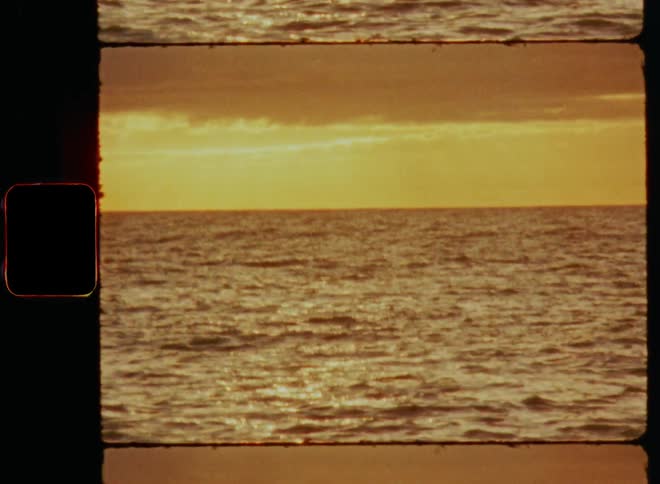 Super 8 footage with tilting upward camera movement of sun that peaks over clouds in red sky onto ocean and beach during sunset with warm golden sunlight and lens flare