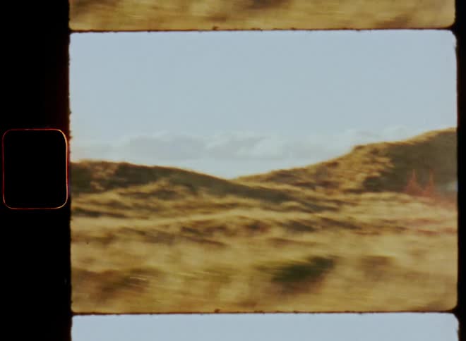 Driving by or passing grassy dunes next to the sea or beach at the coast of the ocean while the hills or hilly landscape is illuminated by bright sunshine on a sunny day with blue sky and cumulus clouds at the horizon