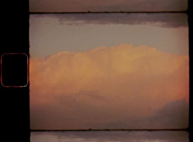 Panning shot of dark clouds that hover above red and colourful cumulus clouds creating a vibrant contrast over the silhouette of a field of grass on Super-8 mm film