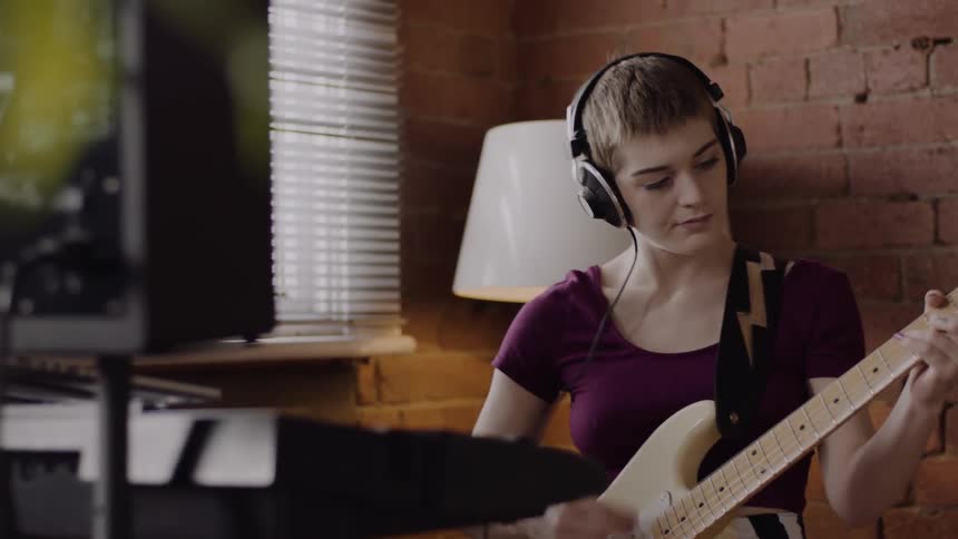 A young woman plays a guitar while standing in a home studio
