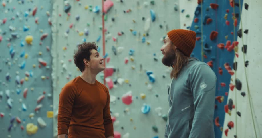 Two men are standing near a climbing wall looking up as they talk
