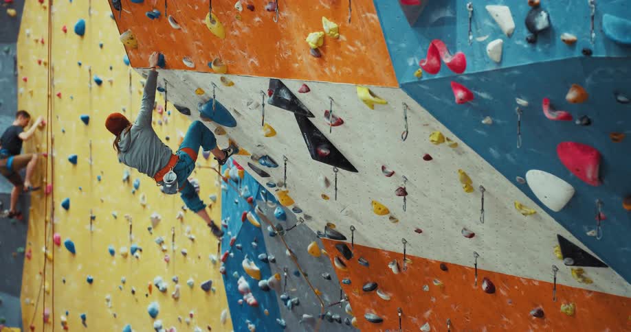 A man climbing a rock wall hangs from one arm and one foothold as he watches another climber