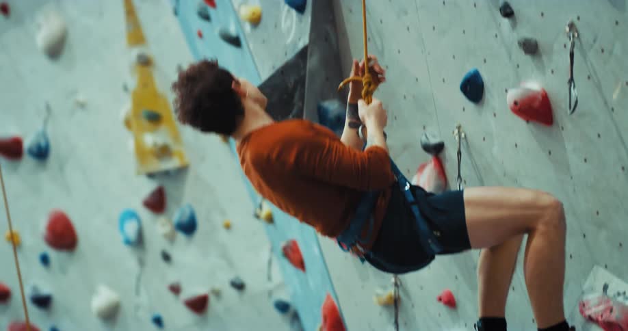 A man has a harness and rope around his waist as he scales a climbing wall and slips as he swings and hangs from his rope