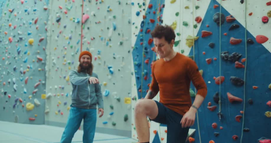 A man demonstrating how to exercise then another man tries to do it in front of a rock climbing wall