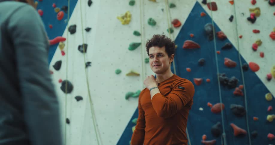 Two men are standing near a climbing wall exercising their arms as they face each other and smile