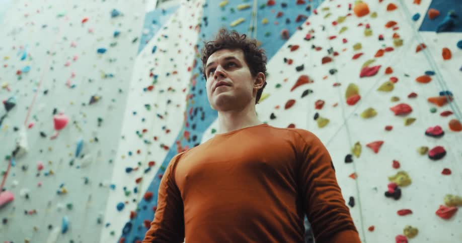 Two men are standing near a climbing wall as one man steps up and looks upward and then steps back down