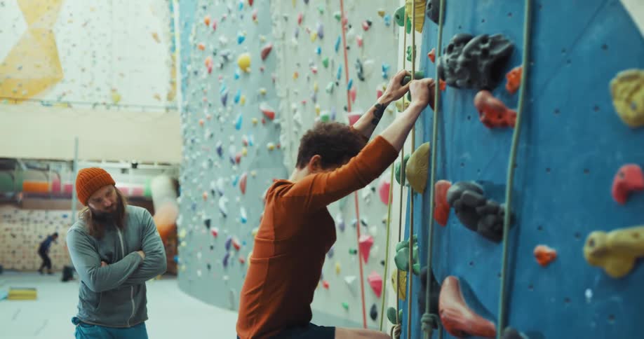 Man stands in climbing gym watching another climb rock wall