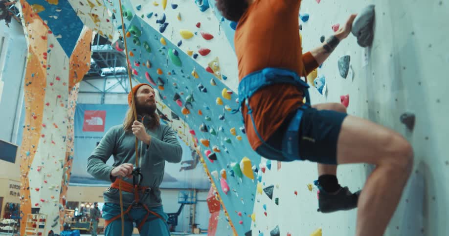 Two male friends practice rock climbing on a colorful interior climbing wall in a gym