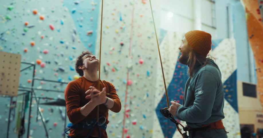 Man holds rope and looks upward in climbing gym before rock climber lowers himself and they celebrate
