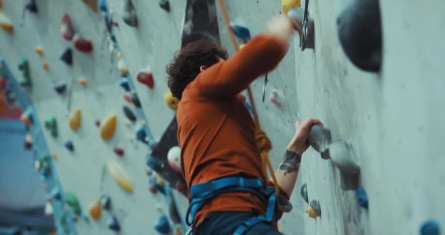 Man wearing harness climbs wall in climbing gym then gestures angrily after slipping to floor