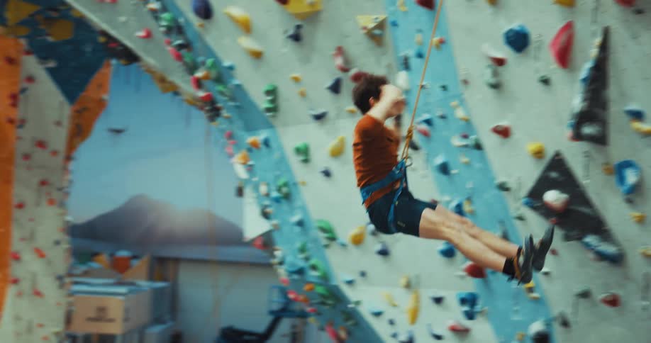 Man wearing harness climbs wall in climbing gym then gestures upset after slipping