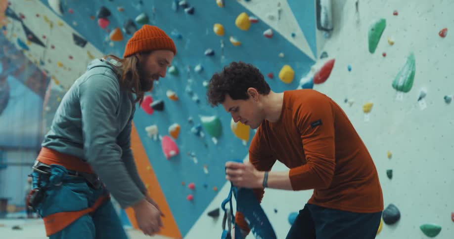 Two men in an indoor climbing center helping each other put on harnesses