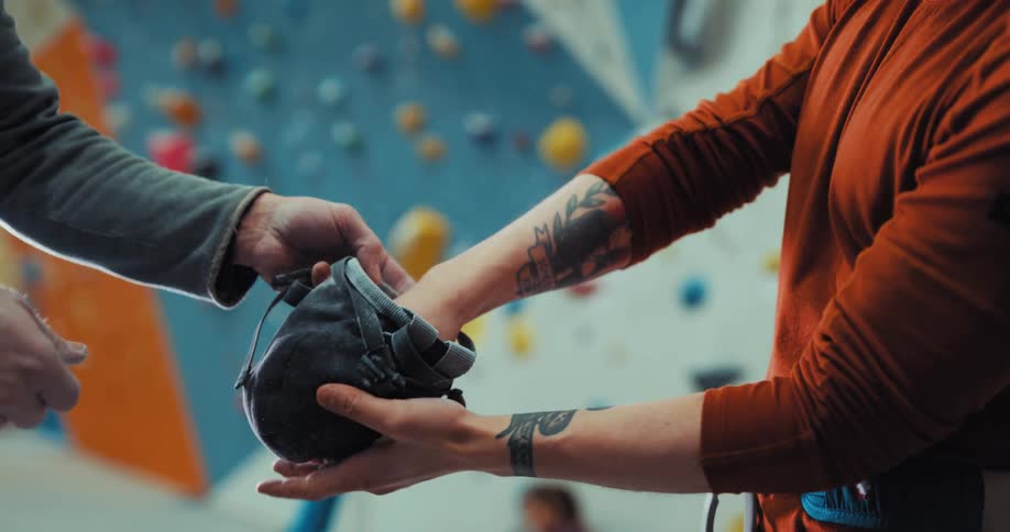 One man holds out bag to help another climber chalk his hands in climbing gym