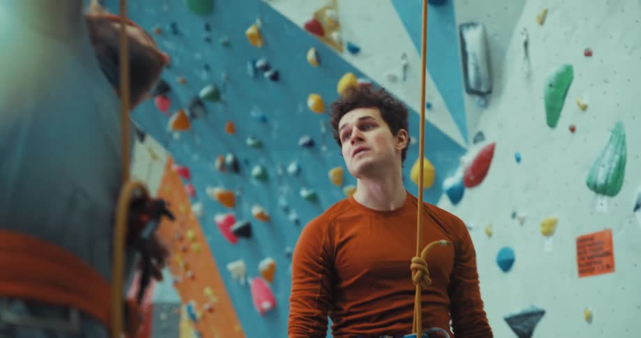 Two young men wearing harnesses in an indoor climbing center discussing strategy to get up the wall