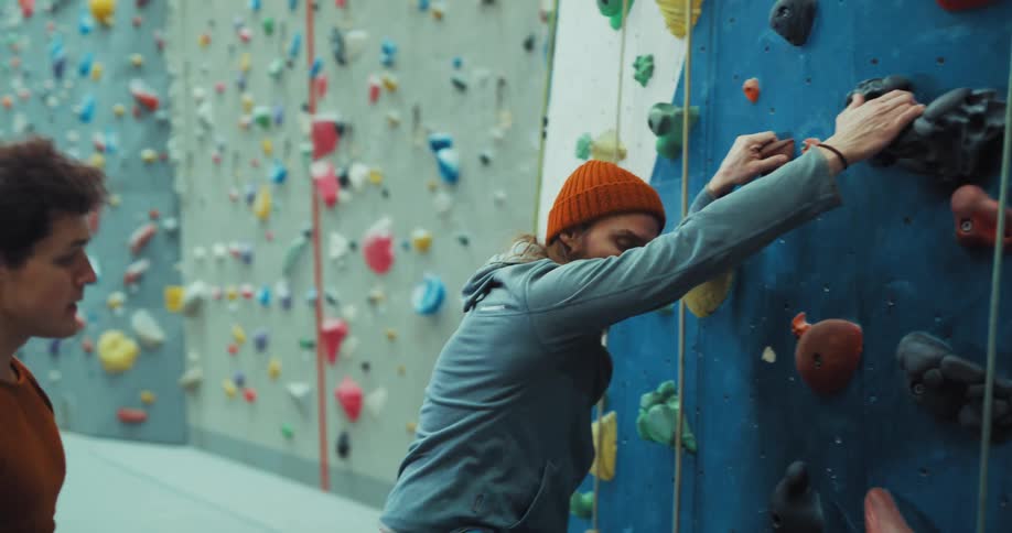 A man trying to climb a rock wall falling off and getting frustrated while the trainer demonstrates for him