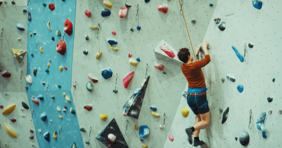 A man has a harness and rope around his waist as he scales a climbing wall and slips as he swings and hangs from his rope