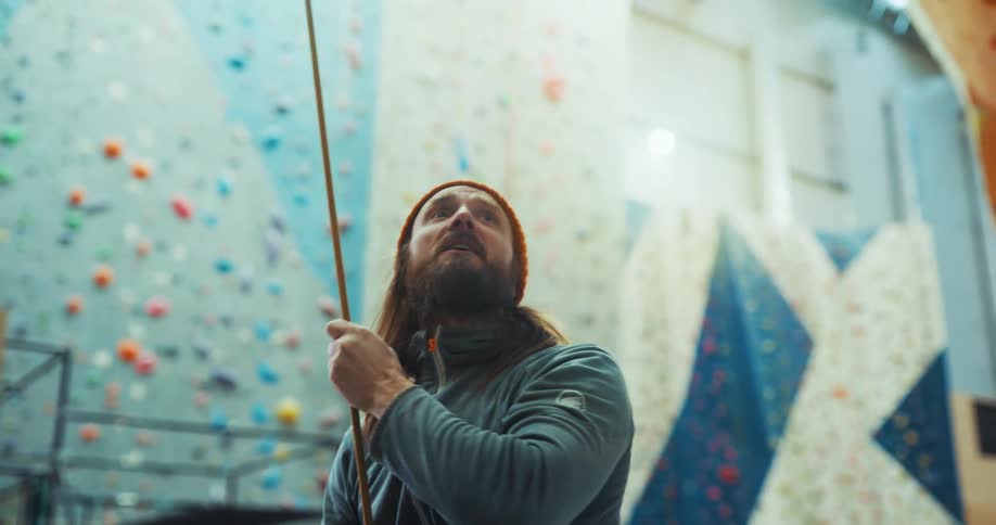 A man is standing near a climbing wall as he holds a rope and points as he speaks to someone