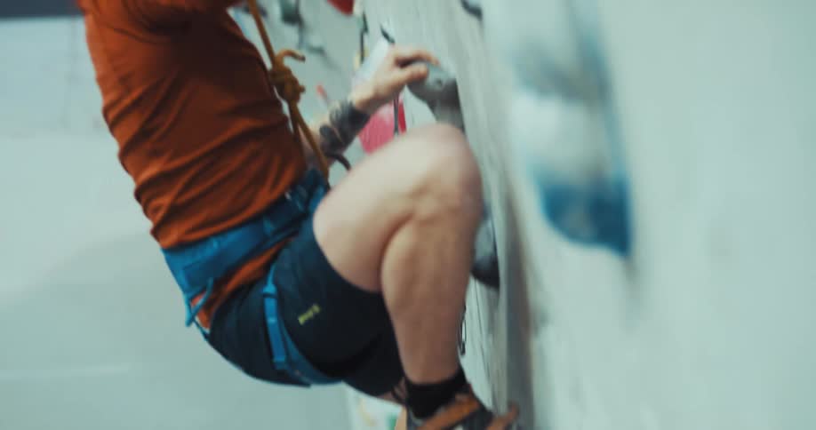 Man holds rope tied to harness of second man climbing wall in climbing gym