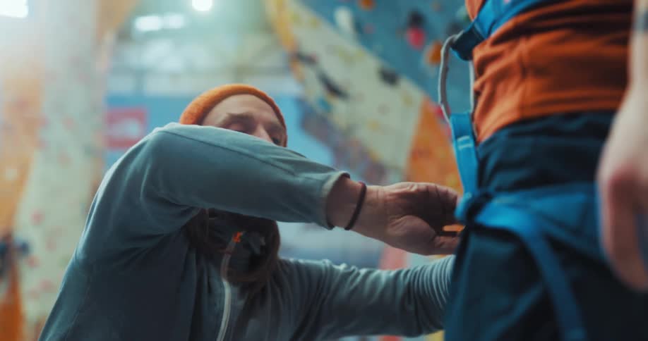Man crouches to adjust harness worn by climber in climbing gym