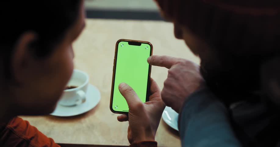 Two men sitting at a table looking at the cellphone in the one mans hands while coffee cups sit on the counter