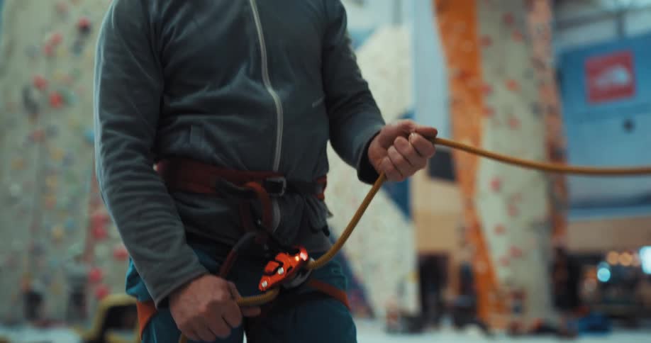 A bearded man adjusts a rope as he stands within an indoor climbing gym