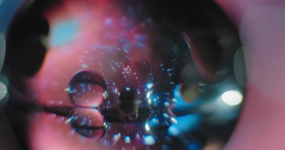 Close up of a large transparent spere that has several smaller spheres embedded in it on a blurry pink and blue background and a light reflecting from the right side