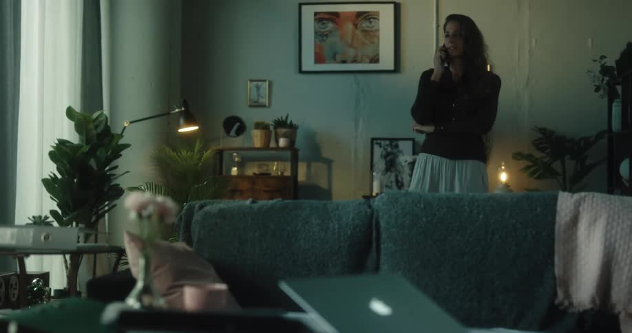 A woman is walking around her living room looking at paperwork in a folder as she talks on her phone