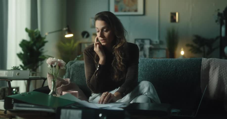 A woman is sitting on her couch talking on her phone and writing on paper and she looks through papers and looks at her laptop computer and leans back looking at her phone