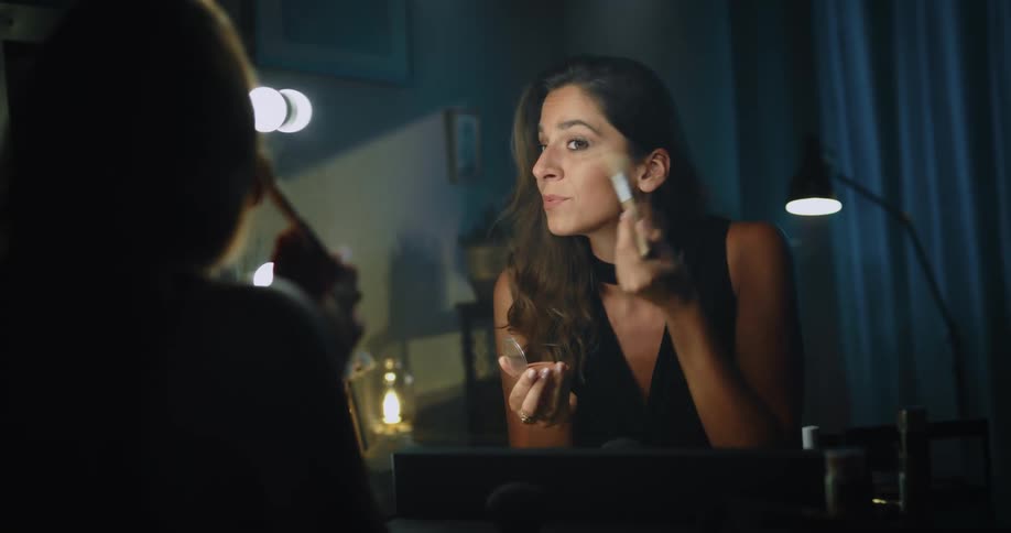 A woman retrieves a makeup brush from a bag and uses it to apply makeup to her face while sitting at a vanity