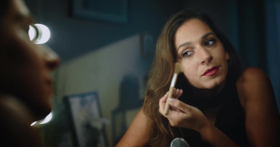 A woman stares into a vanity mirror while applying blush and mascara