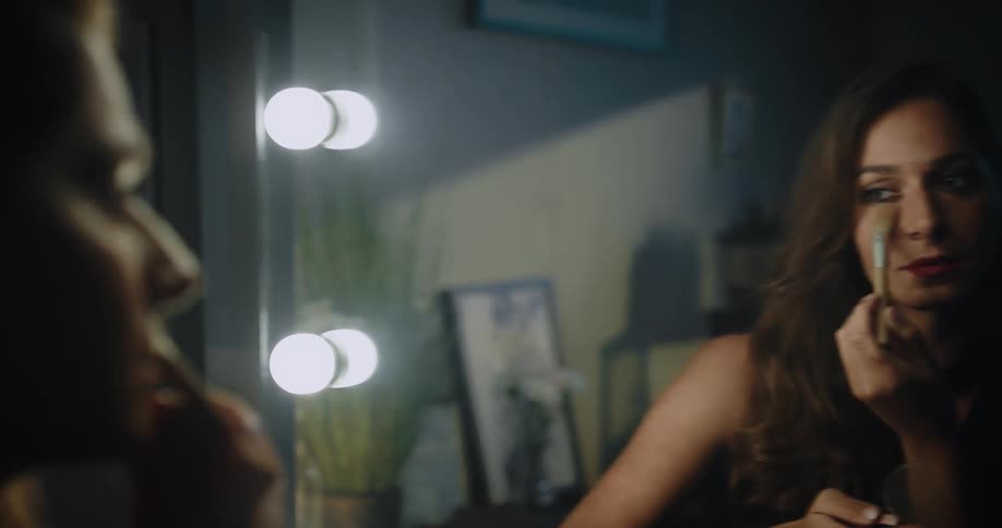 A woman sits in front of a mirror and uses a brush to apply makeup