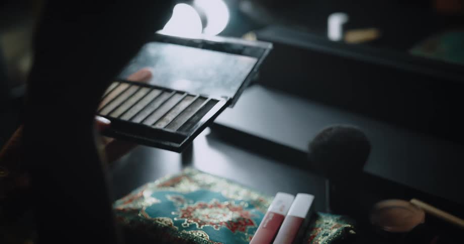 Woman uses small brush to apply makeup while sitting in front of makeup mirror on dressing table