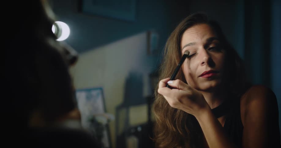 A woman is sitting in front of a mirror looking at her face as she brushes makeup on her eyelids