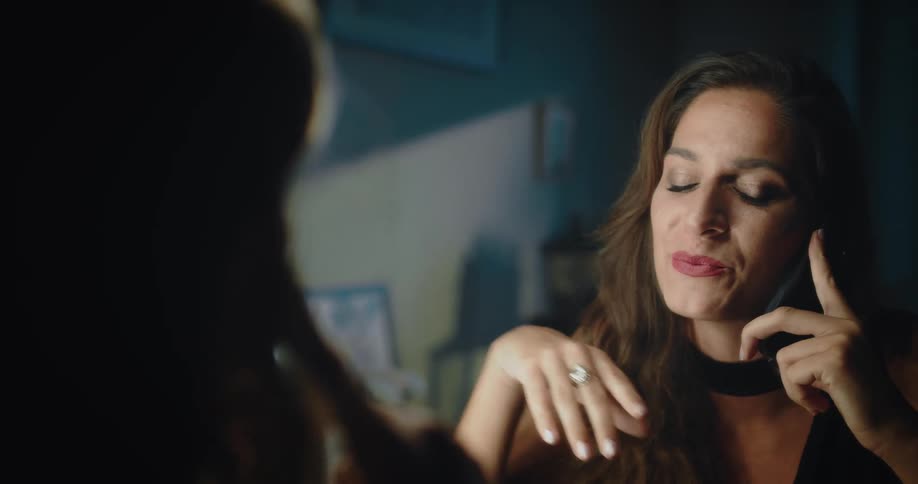 A woman is sitting in front of a mirror and fixes her makeup as she talks on the phone