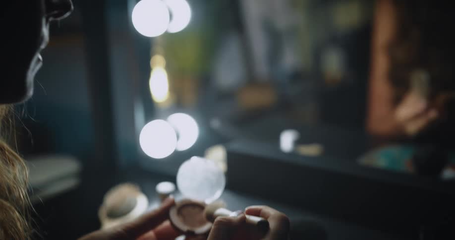 A woman is sitting at a table in front of a mirror as she opens a makeup compact and brushes powder on her face