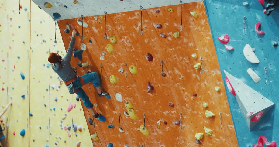 A man is scaling a climbing wall as he reaches for the pouch on his belt to put powder on his hands as he climbs