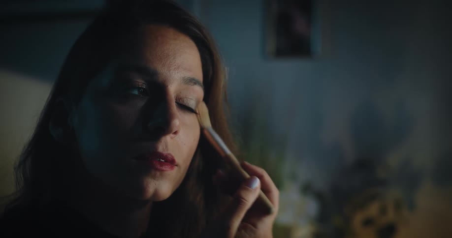 A woman is sitting in front of a mirror looking at her face as she brushes blush on her cheeks