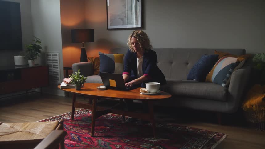 Woman with curly hair puts a mug of coffee on coffee table and goes on to work on laptop in the living room