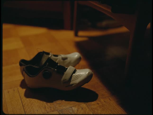A pair of white bicycling shoes rest on a wooden floor the lights turn on and off in the room