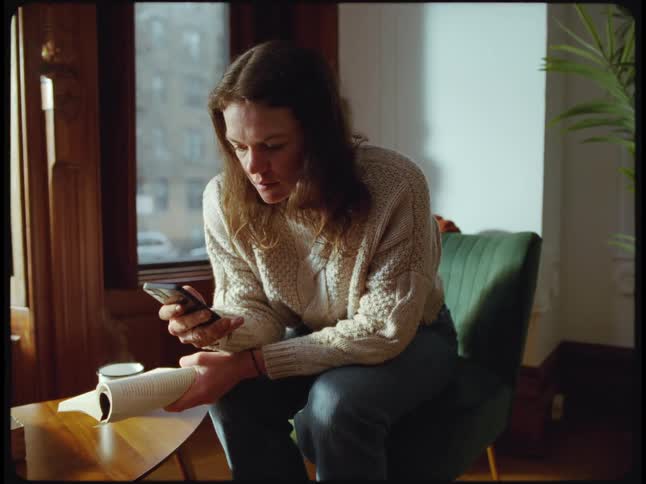 A woman is sitting in a chair holding a book as she uses her cell phone with the other hand