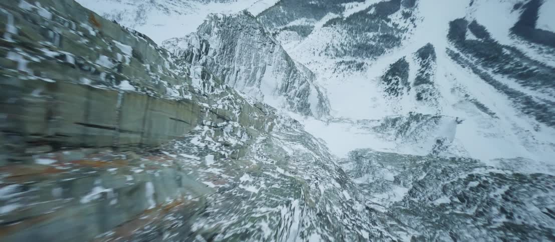 A snow covered mountain peak with snow along the mountainside covering trees and the valley as the wind blows snow through the air