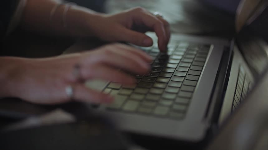 A up close of a persons hand typing on a laptop keyboard with rings on their fingers and a cellphone next to them
