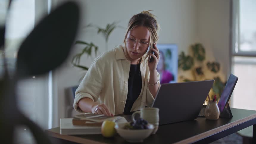 A woman sits at her desk on her cell phone while looking at her laptop
