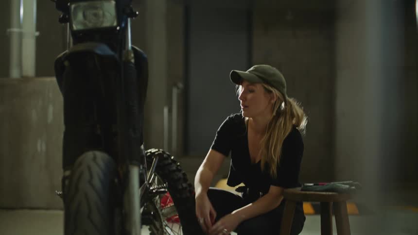 A woman wearing black shirt and pants and a green hat walks over to her motorcycle carrying a tire and kneeling down